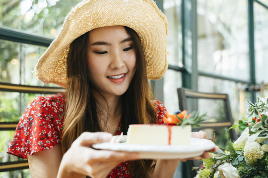 特写镜头：身穿红色连衣裙、戴着帽子、拿着芝士蛋糕盘的泰国亚裔年轻女子。甜点爱好者。