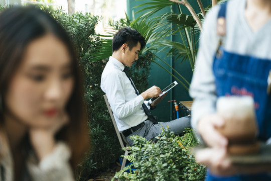 聪明的亚洲商人穿着正装在咖啡厅使用平板电脑。人们聚集在前台。