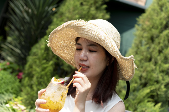 美丽的亚泰女子戴着帽子，在花园里畅饮百香果汁。