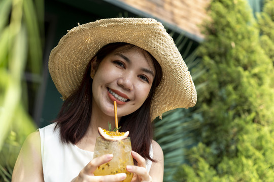 特写镜头：戴着帽子的泰国亚裔年轻女子在大自然中畅饮百香果汁。