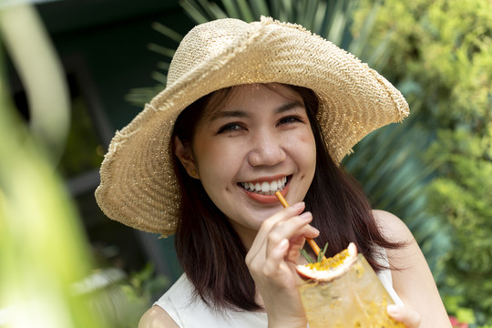 特写镜头：戴着帽子的泰国亚裔年轻女子在大自然中畅饮百香果汁。