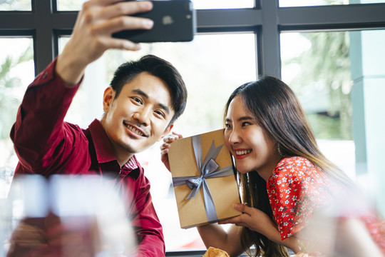 一对身着红色礼服的泰国夫妇给对方一个惊喜。情人节送礼物给女友的男人。浪漫情侣用智能手机自拍，将记忆保