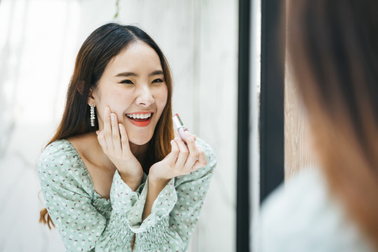 快乐的年轻美丽的亚洲女人穿着绿色时尚衬衫在镜子前涂口红。