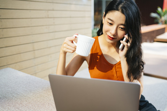 穿着橙色衬衫的性感亚洲女人一边打电话一边喝咖啡一边看着笔记本电脑。