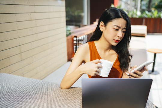 不开心的性感亚洲女人穿着橙色衬衫，一边打电话，一边喝咖啡，一边看着笔记本电脑。