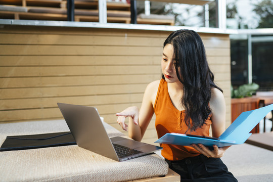性感的亚洲女人穿着橙色衬衫在家里工作拿着蓝色的剪贴板文件看着笔记本电脑。