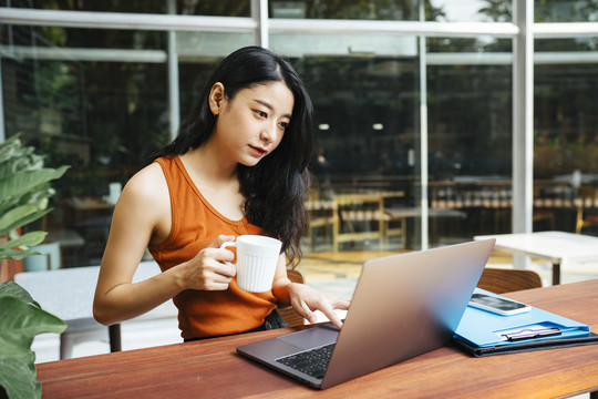 性感的亚洲美女穿着橘色衬衫在咖啡厅用笔记本电脑工作。