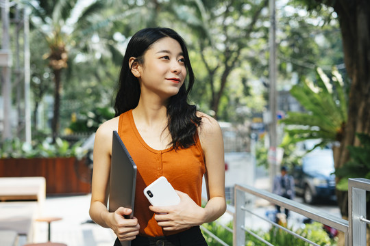 一幅性感的亚洲美女的画像，她穿着橘色衬衫，手里拿着笔记本电脑，在城市的咖啡园里打电话。
