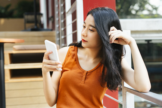 性感的亚洲美女穿着橙色衬衫手持智能手机坐在户外的工作空间。
