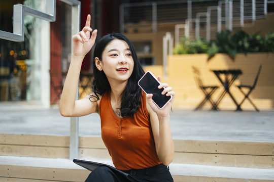 亚洲美女身穿橙色衬衫手持智能手机挥手给朋友打电话。