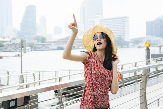 年轻漂亮的泰国亚裔旅行家，穿着红色连衣裙，戴着帽子，戴着太阳镜，指着天空。曼谷市的一位女旅行者。