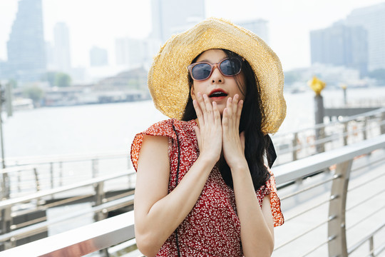 兴奋的年轻美丽的泰国亚裔旅行家妇女的画像，穿着红色的裙子，帽子，戴着太阳镜-惊喜快乐的表情。