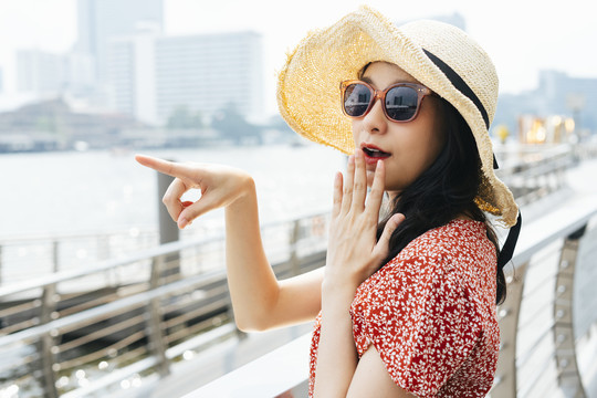 年轻漂亮的泰国亚裔旅行家，穿着红色连衣裙，戴着帽子，戴着太阳镜，指着曼谷河面上的某物。