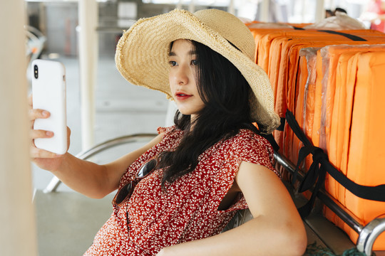 年轻漂亮的泰国亚裔旅行家，穿着红裙子，戴着红帽子，坐在船上。用智能手机和电话自拍。