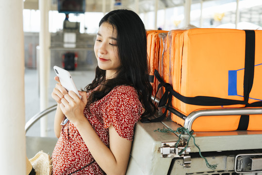年轻漂亮的泰国亚裔旅行家，穿着红色连衣裙，坐在船上用智能手机。