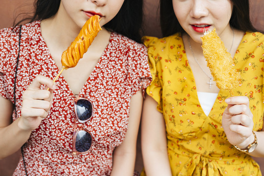两位穿着红黄相间连衣裙的亚洲美女手捧街头美食，对街头美食有一种棍棒式的概念。