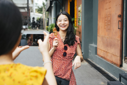 年轻漂亮的亚洲女人穿着红衣服在智能手机镜头前吃着街头食物。美食博客。街头小吃。