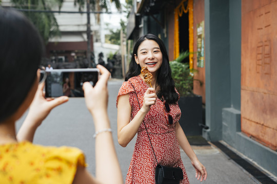 年轻漂亮的亚洲女人穿着红衣服在智能手机镜头前吃着街头食物。美食博客。街头小吃。