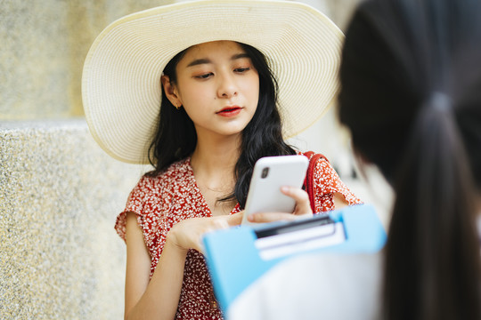 美丽的亚洲女子身穿红色连衣裙，戴着帽子，手持智能手机，神情茫然地走进城市，走进导游工作人员。