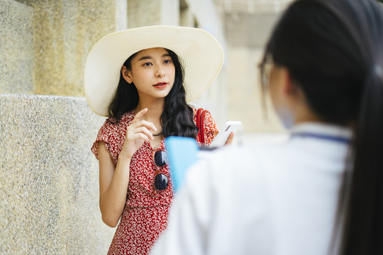美丽的亚洲女游客穿着红色的裙子，戴着红色的帽子，在城市里和导游交谈。