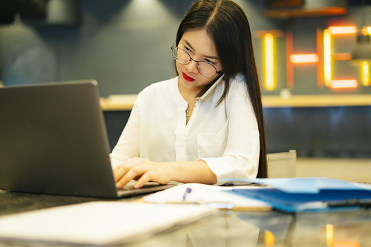 忙碌的亚洲女人戴着眼镜在智能手机上聊天，在笔记本电脑上工作。