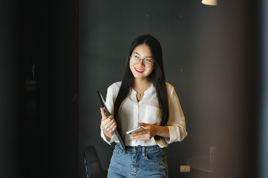 年轻漂亮的亚洲女人戴着眼镜手持笔记本电脑，使用智能手机的肖像。
