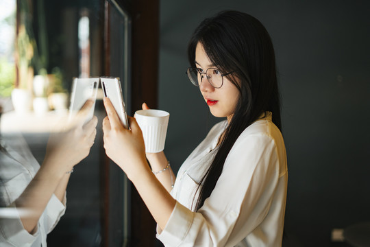 早上，年轻漂亮的亚洲女人戴着眼镜，穿着白色睡衣，手里拿着一杯咖啡，用智能手机靠近窗户。