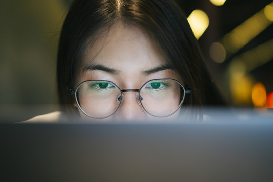 特写镜头：年轻漂亮的亚洲女人戴着眼镜，晚上在笔记本电脑前工作。