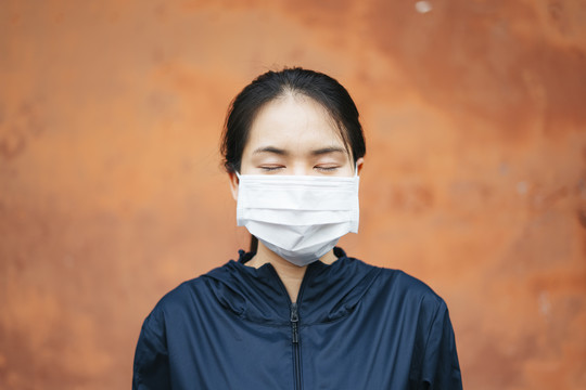 年轻的亚洲女子戴着隔离在橙色背景下的面具，闭上眼睛，烟雾危机重重。