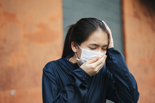 一名戴口罩的亚洲妇女因城市烟雾而咳嗽。双手捂着头。不健康的状况。