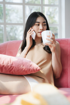 一个长发波浪的女人坐在客厅的粉色沙发上，一边喝着热咖啡，一边和朋友通过电话预约。