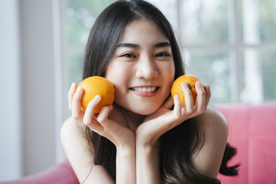 活泼的长发女孩坐在粉色沙发上，手里拿着橘子，笑容灿烂。