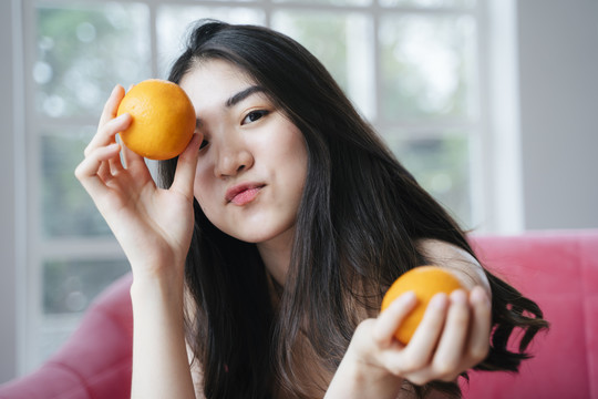 活泼的长发女孩坐在粉红色的沙发上，手里拿着橘子，举着一个橘子在额头，做鸭脸。