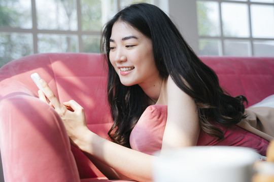 波浪形长发女孩趴在粉色沙发上，一边笑，一边在社交媒体上阅读有趣的内容。