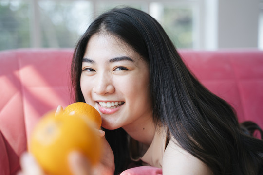 活泼的长发女孩趴在粉红色的沙发上，手里拿着橘子灿烂地笑着，把一个橘子递给镜头。
