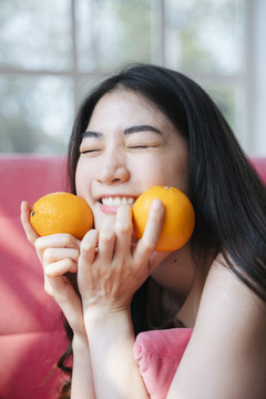 活泼的长发女孩趴在粉色沙发上，手里拿着橘子，笑容灿烂，下巴托在手上。