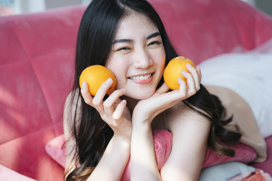 活泼的长发女孩趴在粉色沙发上，手里拿着橘子，笑容灿烂，下巴托在手上。
