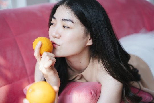 活泼的长发女孩趴在粉色沙发上，手里拿着橘子，亲吻橘子。