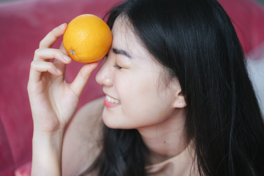 活泼的长发女孩趴在粉色沙发上，手里拿着橘子，笑容灿烂，额头上举着一个橘子。
