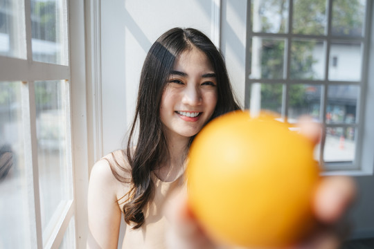 早上，一个长发卷曲的女孩站在窗前，手里拿着橘子，把橘子递给相机。
