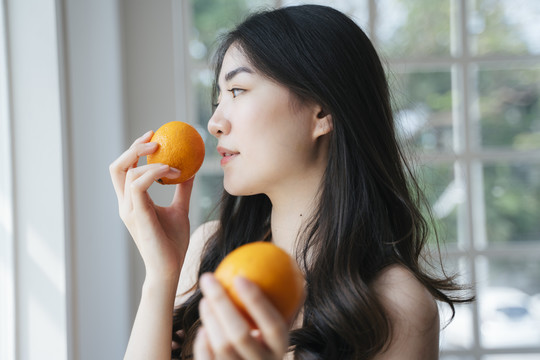 早上，一个长发卷曲的女孩站在窗前，手里拿着橘子，闻到橘子的味道。
