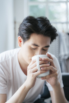 早上，一个穿着白t恤的男人坐在窗前，喝着白杯子里的热咖啡。