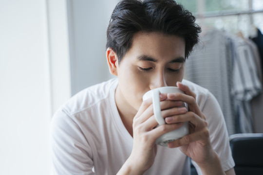 早上，一个穿着白t恤的男人坐在窗前，喝着白杯子里的热咖啡。