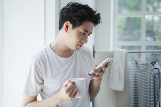 一个穿着白色t恤的男人拿着杯子，一边看手机，一边接听来电。