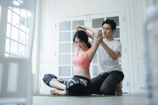 一位穿着旧玫瑰色运动内衣的长发女士在家里接受男性私人教练的瑜伽姿势训练。