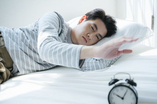 一个男人睡在白色的床上，早上闹钟把他叫醒，他试图阻止它。