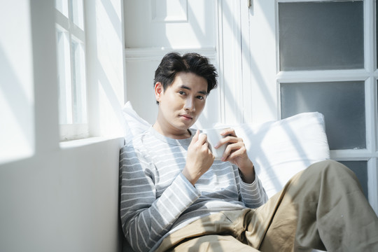 一个男人坐在床上，靠着窗户，端着一杯咖啡，阳光透过窗户。