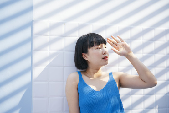 美丽的bob发型亚洲女人，穿着蓝色背心衬衫，在白色瓷砖的背景下遮住了阳光。
