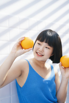 健康胖乎乎的漂亮鲍勃发型亚洲女人穿着蓝色背心衬衫手拿两个橘子水果在白色背景下。