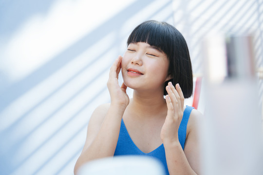 快乐美丽的亚洲女人美丽的鲍勃发型穿着蓝色背心衬衫的亚洲女人在白色房间的脸上涂奶油乳液。